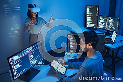 developer uses a VR headset to interact with projected interface designs, while colleagues discuss and code on screens, Stock Photo