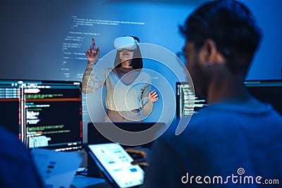 developer uses a VR headset to interact with projected interface designs, while colleagues discuss and code on screens, Stock Photo