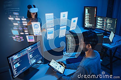 developer uses a VR headset to interact with projected interface designs, while colleagues discuss and code on screens at office Stock Photo