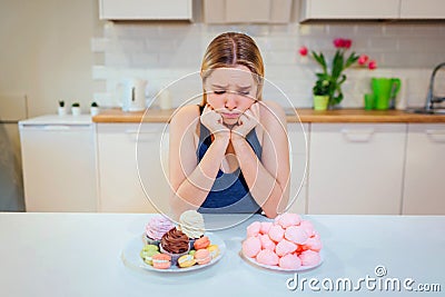 Diet struggle. Young sad woman in blue T-shirt chooses between fresh fruit vegetables or sweets while looking at them in Stock Photo