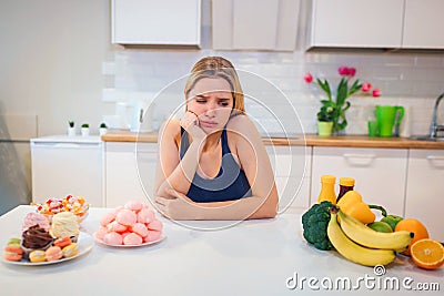 Diet struggle. Young sad woman in blue T-shirt choosing between fresh fruit vegetables or sweets in the kitchen. Choice Stock Photo