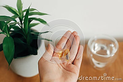 Dietary supplements. Hand holding omega 3 two capsules above wooden table with glass of water. Morning dose of vitamin. Fish oil Stock Photo