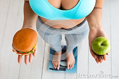 Woman Choosing Between Hamburger And Apple Standing On Weight-Scales, Cropped Stock Photo