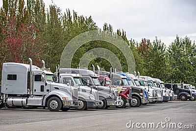 Different big rigs semi trucks standing in row on parking lot with trees on the background Stock Photo