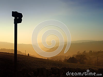 Direction sign in silhouette pointing down a glowing evening sunset valley with golden tree covered hills at twilight Stock Photo