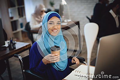 Disabled arab woman in wheelchair working in office. Woman is working on desktop computer and drinking coffee. Stock Photo