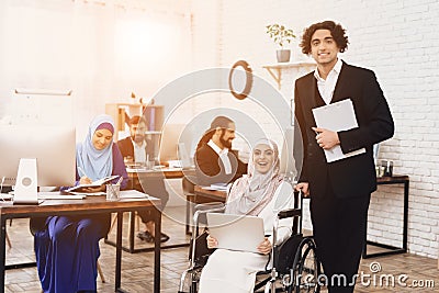 Disabled arab woman in wheelchair working in office. Woman is posing with male coworker. Stock Photo