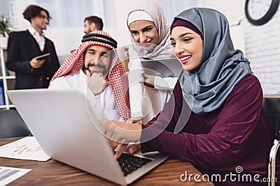 Disabled arab woman in wheelchair working in office. Woman is showing notes on laptop. Stock Photo