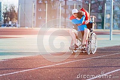 Disabled athlete in a wheelchair on an athletics track. Son hugs his father. Stock Photo