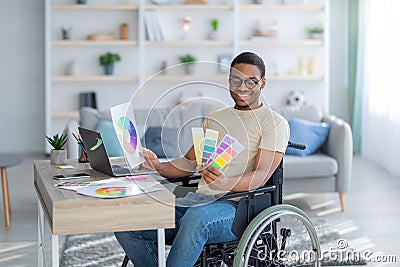 Disabled black guy in wheelchair working as graphic designer online, holding color swatches palette, using laptop indoor Stock Photo