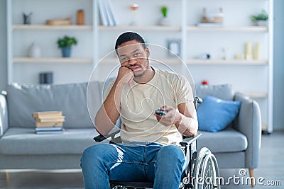 Disabled black man in wheelchair using remote control, watching dull movie on TV, cannot find interesting program Stock Photo