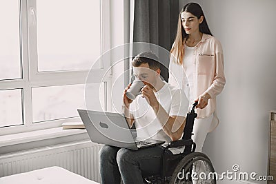 Young man in wheelchair using laptop and girlfriend hugging him at home Stock Photo