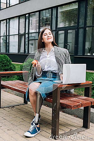 Disabled woman with apple sitting on bench and using laptop on street Stock Photo