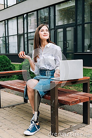 Disabled woman with apple sitting on bench and using laptop on street Stock Photo