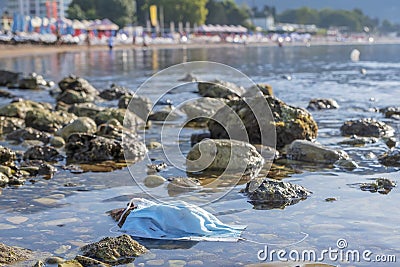 Discarded used facial mask lies on a pebble beach, in the background a beautiful beach with sun loungers and beach parasols. Budva Stock Photo