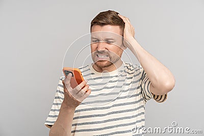 Disgruntled sad shocked frustrated man looks at smartphone screen touches head on grey background. Stock Photo