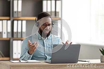 Disgruntled shocked sad young african american bearded male in glasses, headphones gestures and works on pc Stock Photo