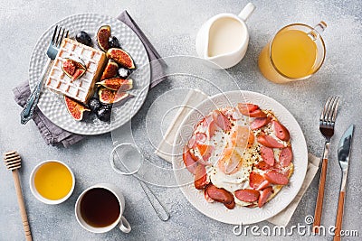 Dishes for home cooked hearty Breakfast. Fried eggs with sausages and tomatoes. Belgian fluted waffles with figs and grapes. Gray Stock Photo