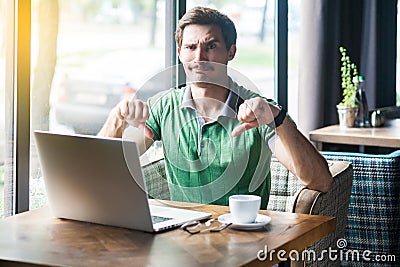 Dislike! Young dissatisfied businessman in green t-shirt sitting, working on laptop, looking at camera and showing thumbs down Stock Photo