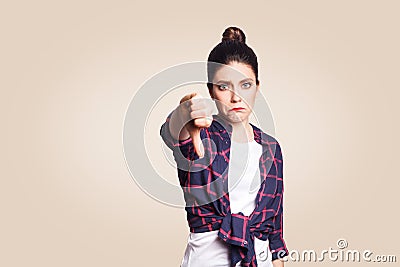 Dislike. Young unhappy upset girl with casual style and bun hair thumbs down her finger, on beige blank wall with copy space looki Stock Photo