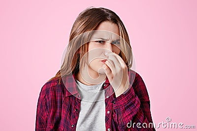 Displeased female model plugs nose as smells unpleasant scent, something disgusting and stinky, has unhappy and irritaed look, iso Stock Photo
