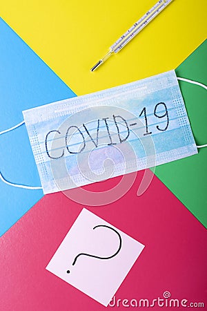 Disposable medical mask with COVID-19 written on it, thermometer and sticker with question mark against the multicolored geometric Stock Photo