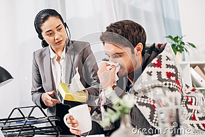 Disquieted call center operator giving napkins to sick coworker with blanket and cup Stock Photo