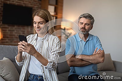 Dissatisfied adult caucasian husband looks at wife with smartphone, lady suffers from gadget addiction Stock Photo