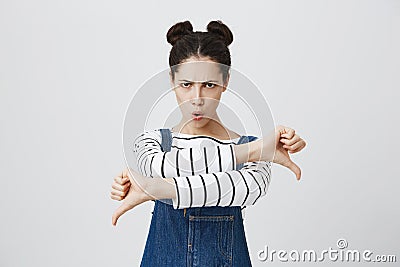 Dissatisfied angry furious girl with hairbuns in denim overalls giving two thumbs down gesture, expressing her Stock Photo