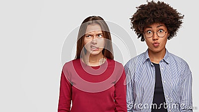 Dissatisfied European woman frowns face, clenches teeth, looks in displeasure, stands near her dark skinned female Stock Photo