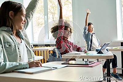 Diverse schoolkids sitting at desk raising hands answering at lesson. Stock Photo