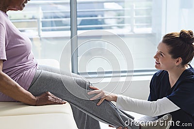 Diverse senior female patient exercising leg and female doctor advising in hospital room Stock Photo