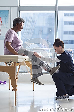 Diverse senior female patient exercising leg and female doctor advising in hospital room Stock Photo