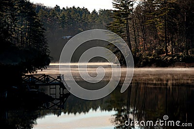 A dock reflects in calm lake water in a misty sunrise in Western Stock Photo