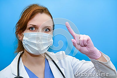 The doctor makes a careful finger - up gesture, close-up. A woman in medical protective gloves and a mask on a blue background, Stock Photo