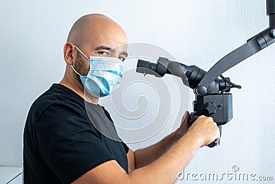 Doctor in medical mask holding handles of black dental microscope and looking aside while working in odontic hospital. Stock Photo