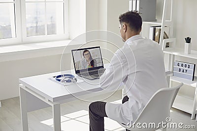 Doctor online. Male doctor works talking video call with patient using bast shoe while sitting at desk in back view Stock Photo