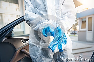Doctor, scientist or nurse wearing face mask and biological hazmat protective suit putting on rubber gloves near car outdoor. Stock Photo
