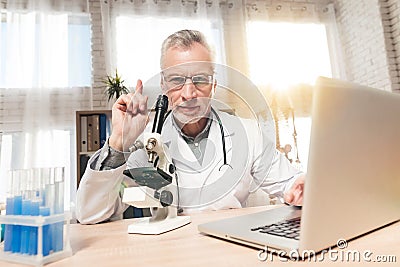 Doctor sitting at desk in office with microscope and stethoscope. Man is looking in microscope. Stock Photo