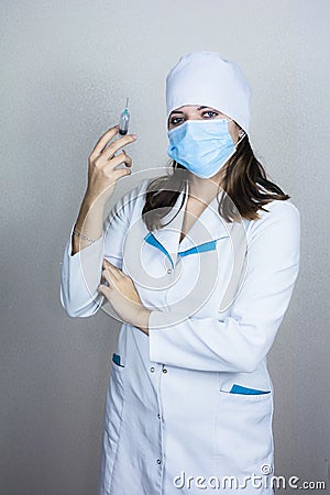 Doctor in white coat with dark hair in medical blue mask holds syringe in hands on light background Stock Photo