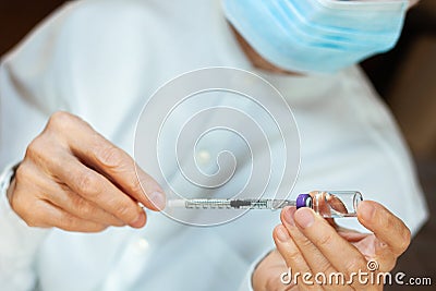 Doctor in a white surgical gown and blue mask holds in hands a medical syringe and bottle with insulin for diabetes. Sugar Stock Photo