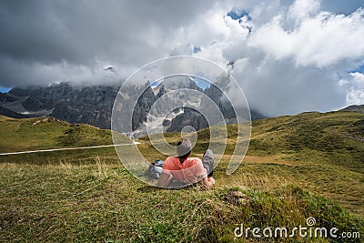 Dolomites. Man laying on the grass enjoying Baita Segantini mountain with Cimon della Pala peak, refuge and lake in Stock Photo