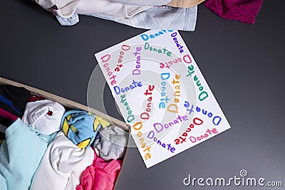`Donate` sign handwritten with multicoloured letters. A box with clothes and a pile of clothes nearby on a grey table Stock Photo
