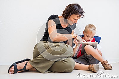 A down-syndrome school boy sitting on the floor with teacher, using tablet. Stock Photo