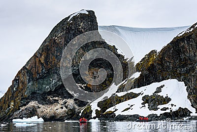 Dramatic Antarctic snow covered and rocky shoreline in Paradise Harbor, Antarctica, with small inflatable rafts cruising in foregr Stock Photo