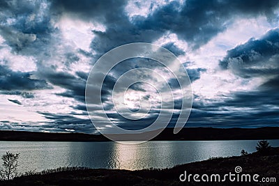 Dramatic dark sky with deep blue clouds and pale yellow sun rays above the lake and mountains covered in shadows. Stock Photo