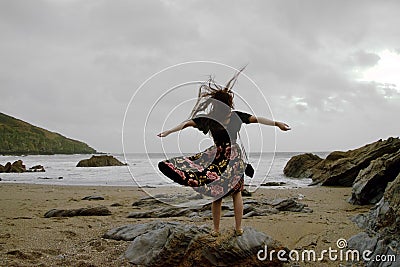 Dramatic portrait of long haired lady in floral formal dress on a stormy beach with arms open Stock Photo