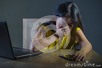 Dramatic portrait scared and stressed Asian Korean teen girl or young woman with laptop computer and mobile phone suffering cyber Stock Photo