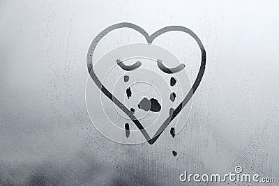 drawn sad face in shape of heart with closed eyes and tears on foggy glass window raindrops blue color concept photo Stock Photo