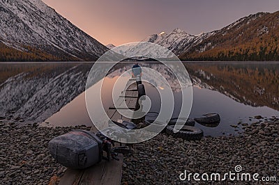 Dreamer. Lonely Man Greets The Dawn Sitting On A Boat Dock On The Shore Of Mountain Lake. Sunrise In Mountains. Old Boat Motor On Stock Photo